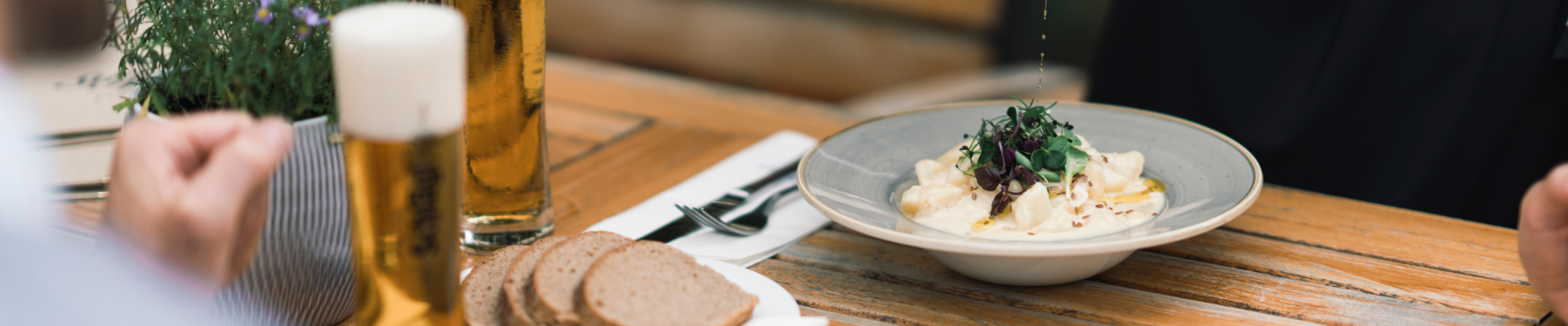 Eine Frau und ein Mann sitzen im Gastgarten des Stift Schlägl und essen Mühlviertler Leinöl Erdäpfel begleitet von ein Paar Scheiben Brot und einem Glas Bier aus der Brauerei Schlägl. Die Frau deren Oberkörper zu sehen ist, ist gerade dabei ihr Gericht mit Leinöl zu garnieren, den Mann sieht man nur undeutlich am linken Bildrand.